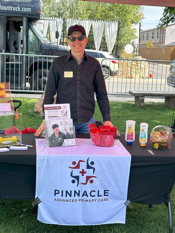 Man at Pinnacle Advanced Primary Care booth; smiling, wearing hat and sunglasses. Outdoor setting with black table.