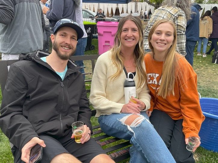 Three friends smiling, outdoors; woman in burnt orange sweatshirt, others holding drinks.