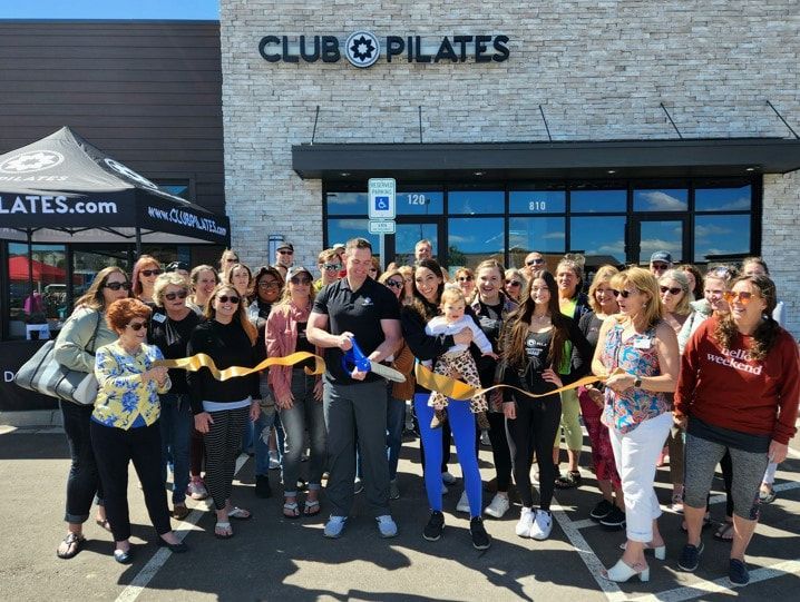 Group cuts ribbon at Club Pilates grand opening, exterior shot. Many smiling, sunny day.