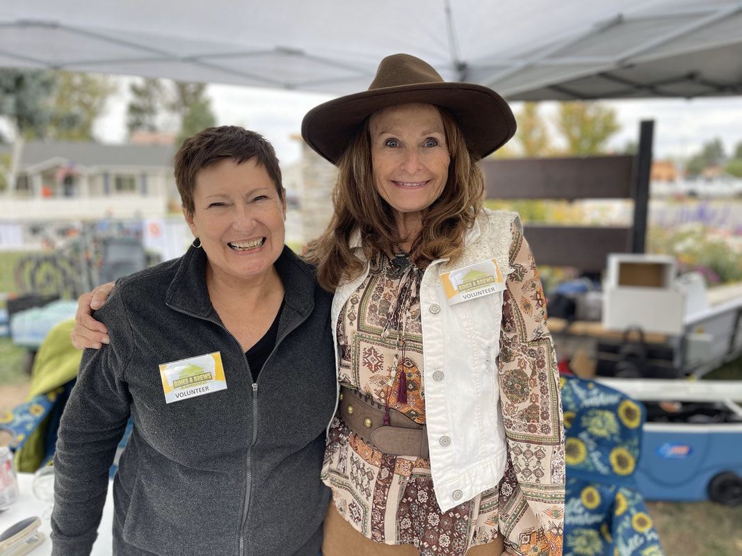 Two women smile, posing outdoors at a market, one with a hat and vest, the other with a fleece jacket.