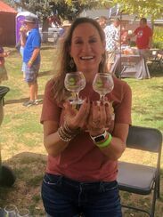 Woman smiles, holding two beer glasses outdoors. She wears a red shirt, jeans, and bracelets.