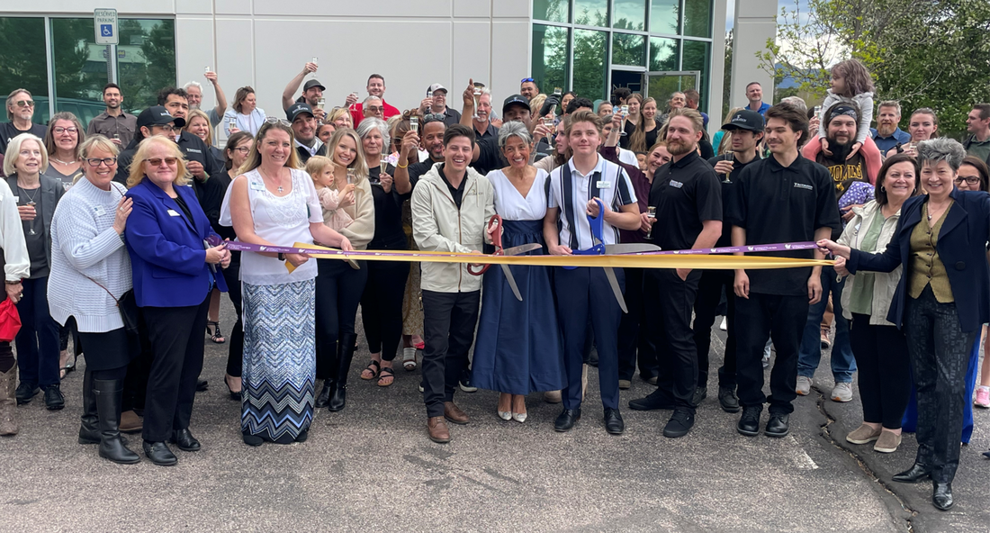 Group of people cutting a ribbon at a building's opening, celebrating. Many smiling faces, diverse ages, and some are raising hands.