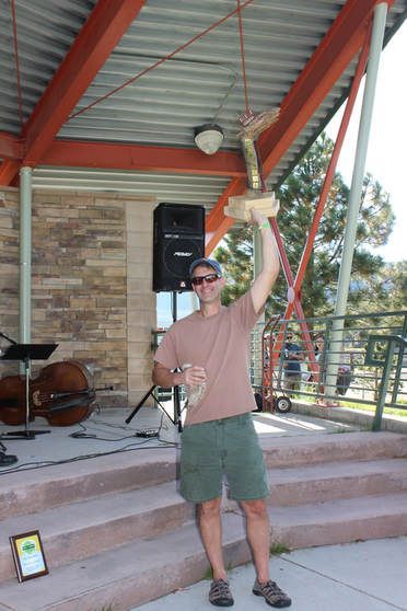 Man holding trophy aloft on stage, brown shirt, green shorts. Outdoor setting, music equipment.
