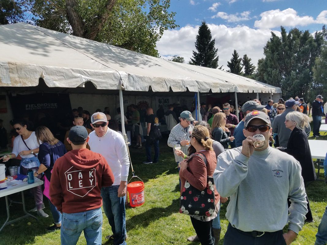 People gathered at a tented outdoor event on a sunny day. Many are socializing, some eating or drinking.