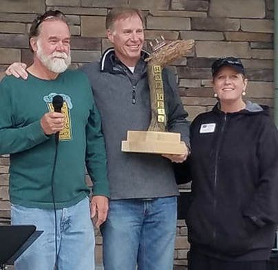 Three people at an outdoor event: man holding award flanked by two others, one with a microphone.