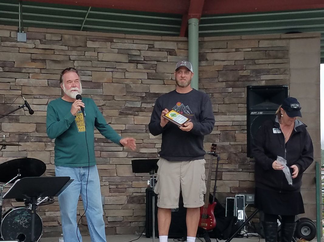 Three people on a stage: a man speaking into a mic, a man holding a gift, and a woman holding a paper.