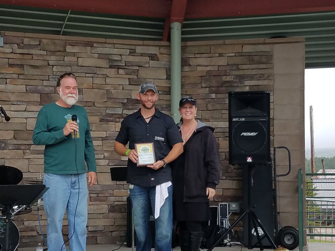 Three people on stage, man holding award, stone wall backdrop, outdoor event.
