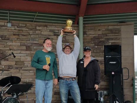 Man holding trophy high, flanked by two people on a stage, celebrating. Stone wall backdrop, speaker and microphone.