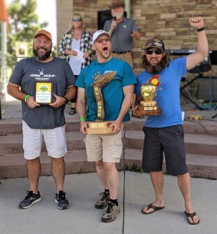 Three men holding trophies, smiling, and celebrating a win on a stage, outdoors.