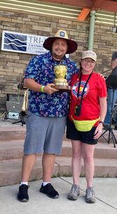 Man in hat and floral shirt holds trophy with woman in red shirt; outdoor event.