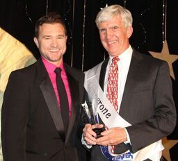 Two men in suits at an event. One man holds an award and sash that reads