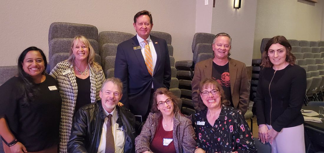 Group of nine people, smiling, posing indoors. Some standing behind others who are seated. Neutral background.