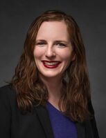 Woman with red hair smiling, wearing a black blazer over a blue shirt.