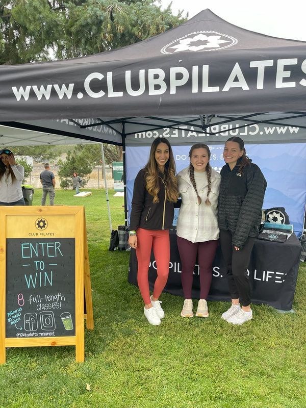 Three women at a Club Pilates booth; one with sign 