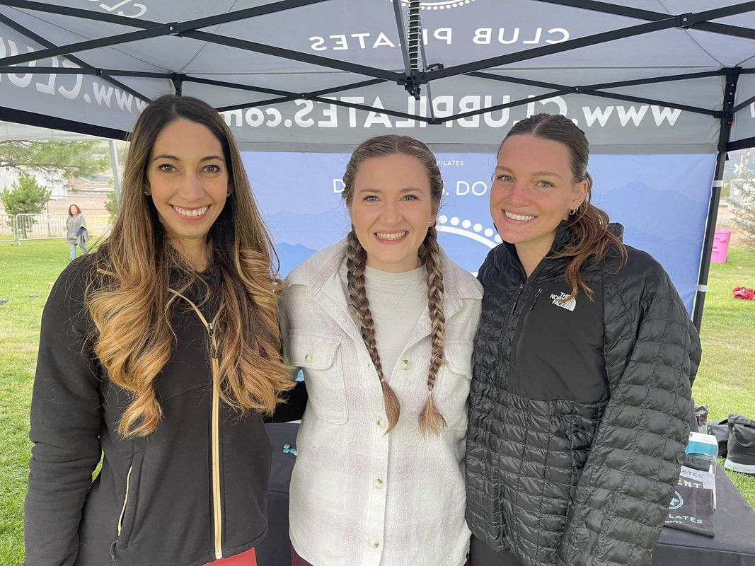Three women smile, posed in front of a banner.  One wears a black jacket, one a white sweater and braids, and one a black jacket with a ponytail.