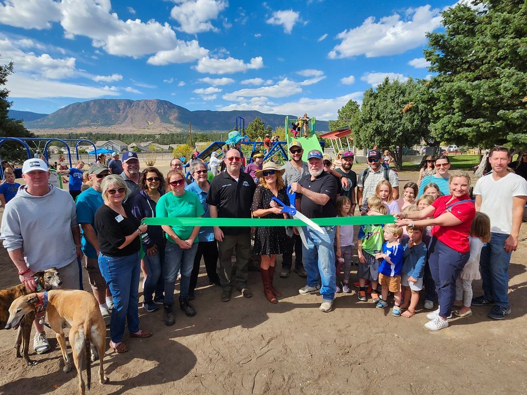 People cutting a green ribbon at a playground opening. Mountains and blue sky are in the background.
