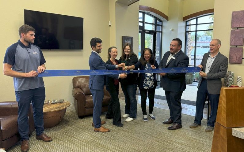 People cutting a blue ribbon at an office grand opening. Men in suits, others in casual wear, pose.
