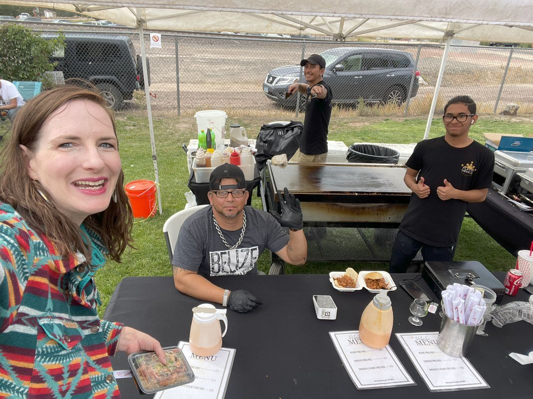 A woman smiles next to a food vendor's table. Three men work at the grill, under a tent.