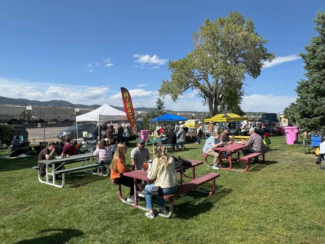 People sitting at picnic tables in a park, eating food with food trucks and a blue sky in the background.