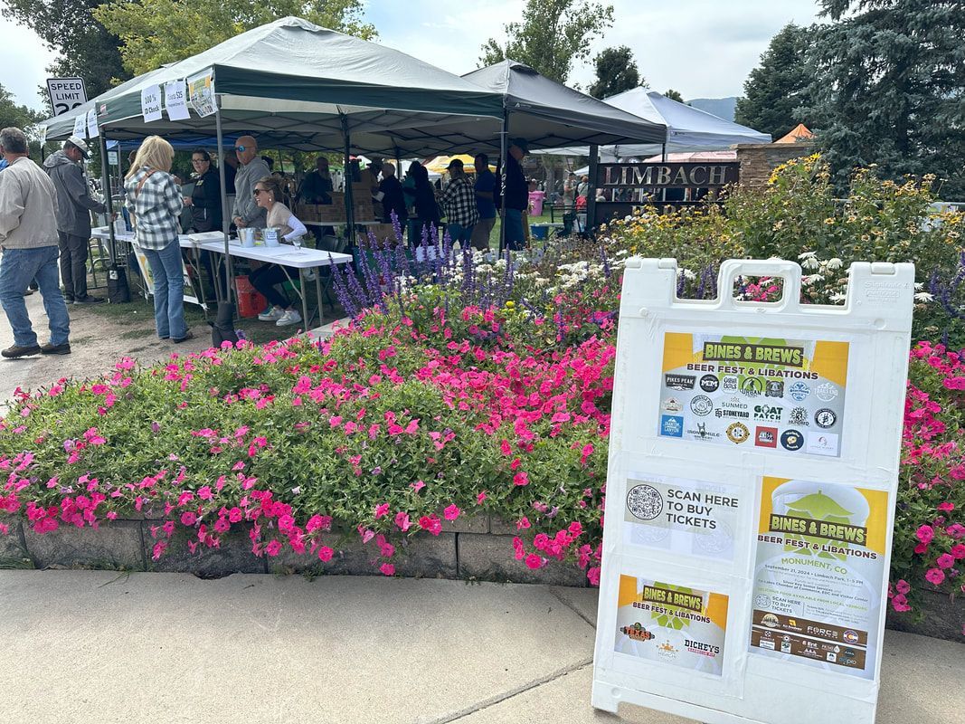 Outdoor farmers market with vendors under tents. People browsing and a sandwich board sign. Pink flowers in foreground.