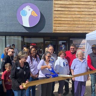 Ribbon-cutting at a business with a goose logo, people smiling, wearing purple.