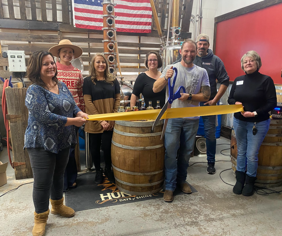 Group cuts yellow ribbon at a distillery grand opening with barrel and flag in the background.