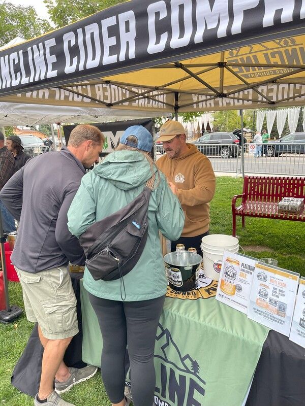 People at a cider booth. A man in a brown hoodie serves customers under a tent with the company name.