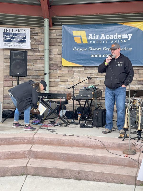 Man speaks into a microphone on stage, woman adjusts equipment. Banner: Air Academy Credit Union.