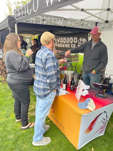 People at a wine tasting: Man pouring wine, others sampling, vendor booth, green grass setting.