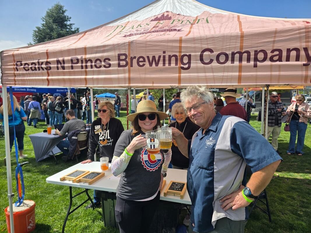 People at Peaks N Pines Brewing Company tent; two smiling, holding beer. Outdoor event, sunny day.
