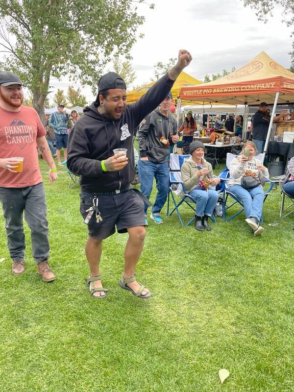 Man raises fist at an outdoor beer festival. He wears black shorts and sandals. People sit at tables in the background.
