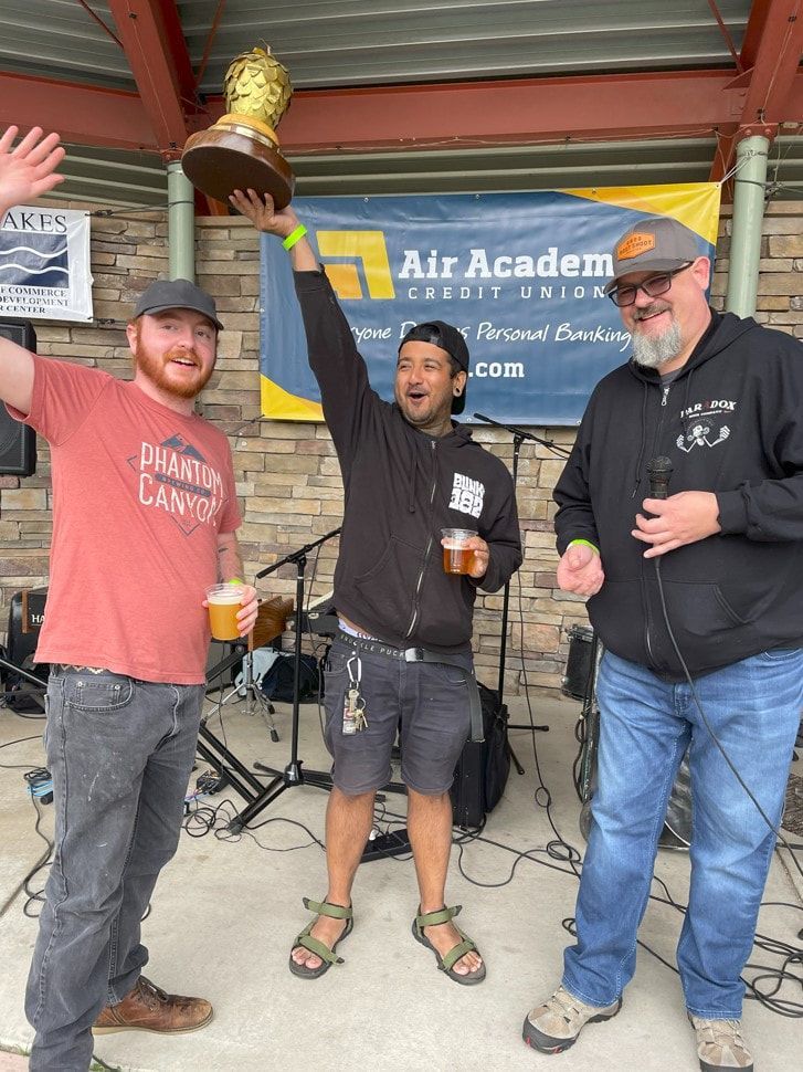 Three men celebrate with a trophy outdoors; one raises it, others hold beer. Air Academy banner.