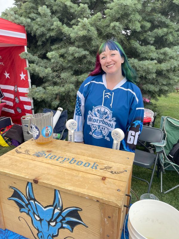 Woman in blue jersey with colorful hair, smiling behind a wooden bar with a dragon logo. Outdoors.