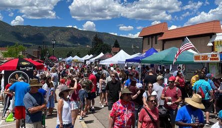 Street fair with many people, tents, and buildings under a blue sky with mountains in the background.