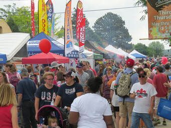 Crowd at outdoor festival with vendor tents, colorful flags, and people walking.
