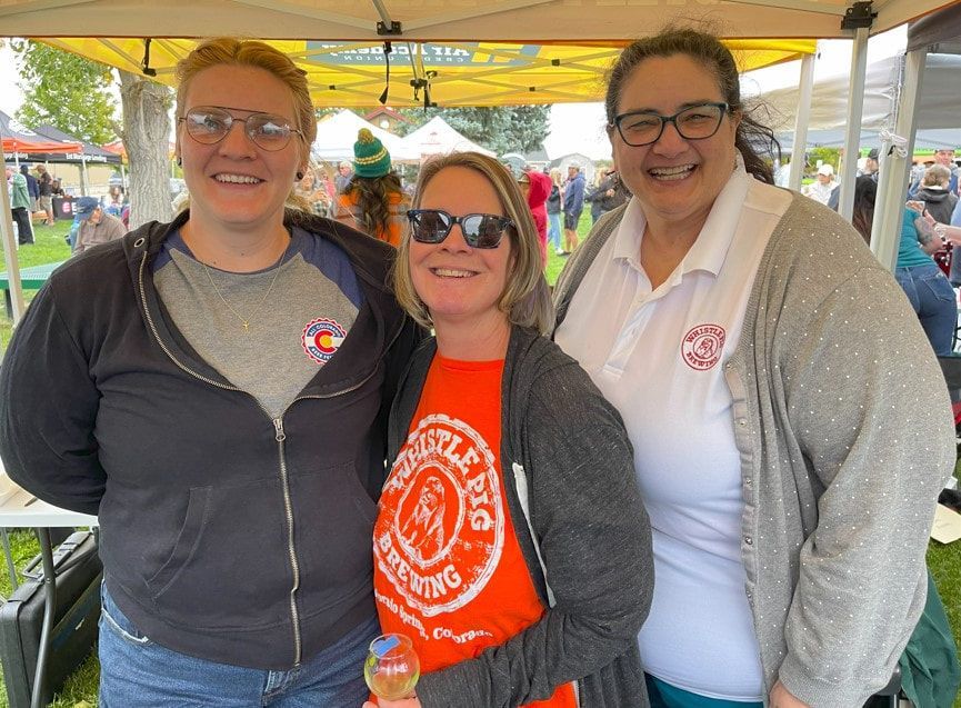 Three women smiling at an outdoor event; one in orange shirt with sunglasses.