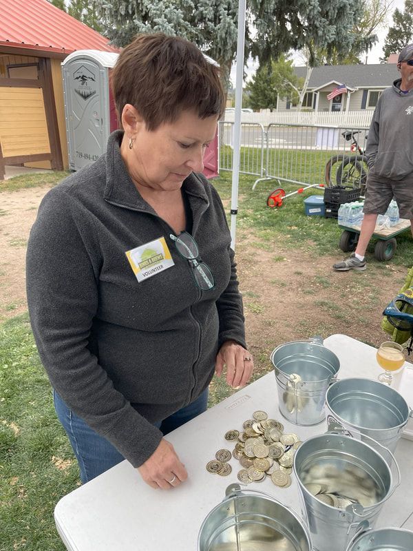 Woman sorting stones at a table, four metal buckets sit nearby. Outdoor setting.