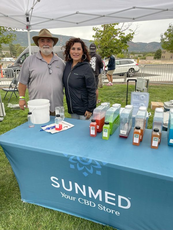 Two people at a SUNMED CBD booth; products displayed on a table. Outdoor setting.