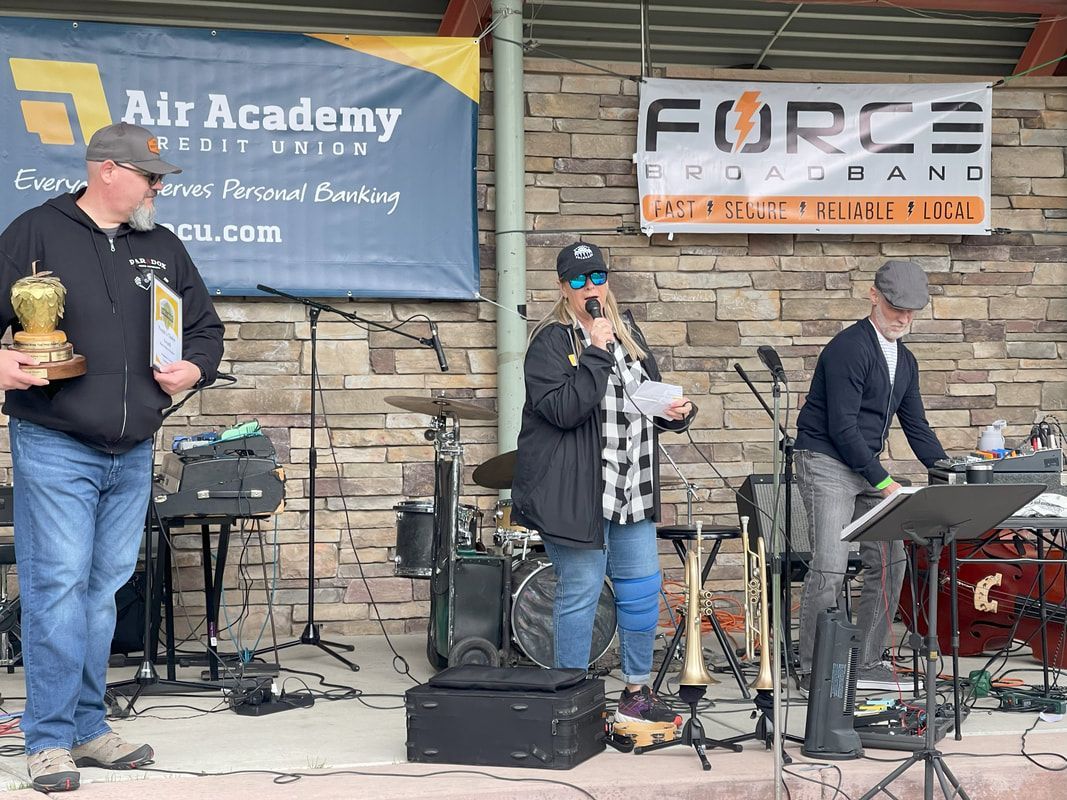 A band performs outdoors. Three people stand on a stage with instruments. One person speaks into a microphone.