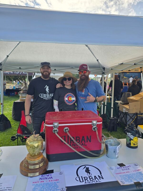 Three people pose at an outdoor booth for Urban Animal. They are by a red cooler and award, smiles.