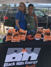 Two women at a Black Hills Energy booth, with bags, a bowl of candy, and a black table cover.