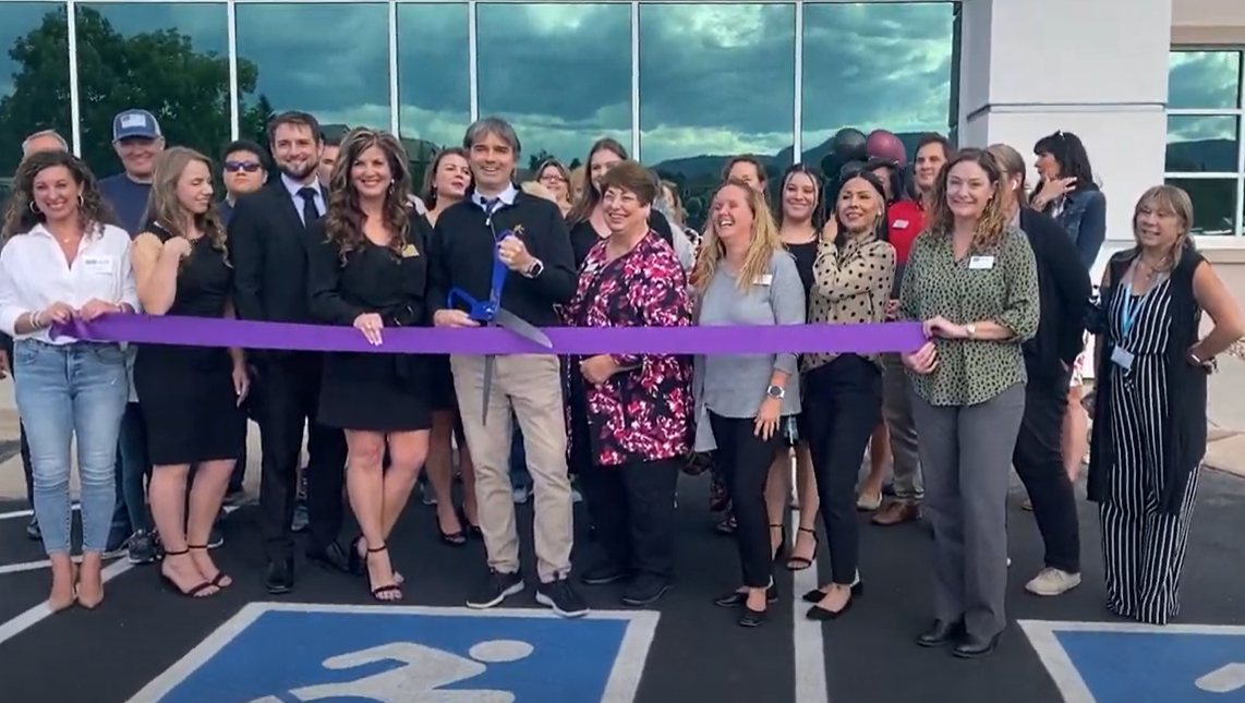 People cutting a purple ribbon at a grand opening. Group of diverse people smiling in front of building.