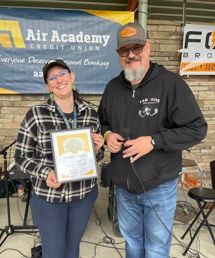 Woman in plaid holding award, smiles at man. Outdoors, Air Academy Credit Union banner.