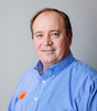 Man in blue shirt with orange logo smiles at the camera.