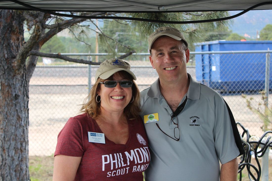 Woman and man smiling, standing under a shade. The woman wears a maroon shirt. The man has a gray polo and a hat.