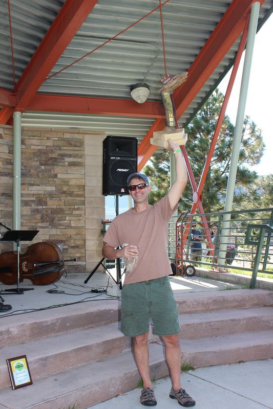 Man in sunglasses holds up trophy, standing onstage outdoors, brown shirt, green shorts.