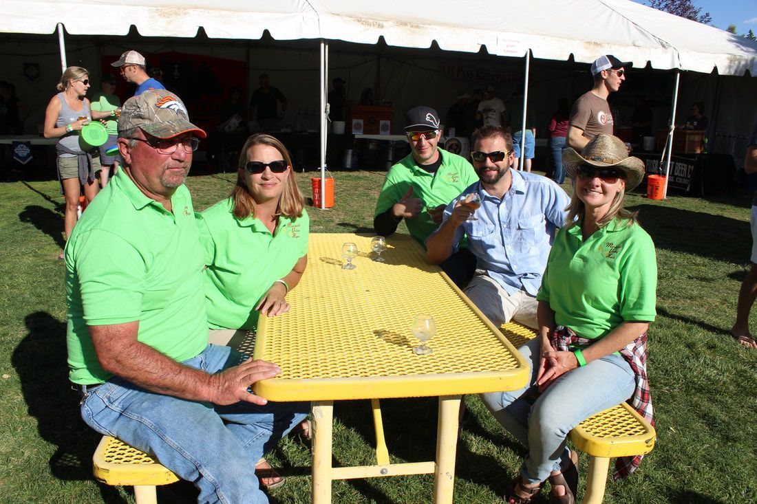 A group of people in green shirts sit at a yellow picnic table under a tent, smiling.