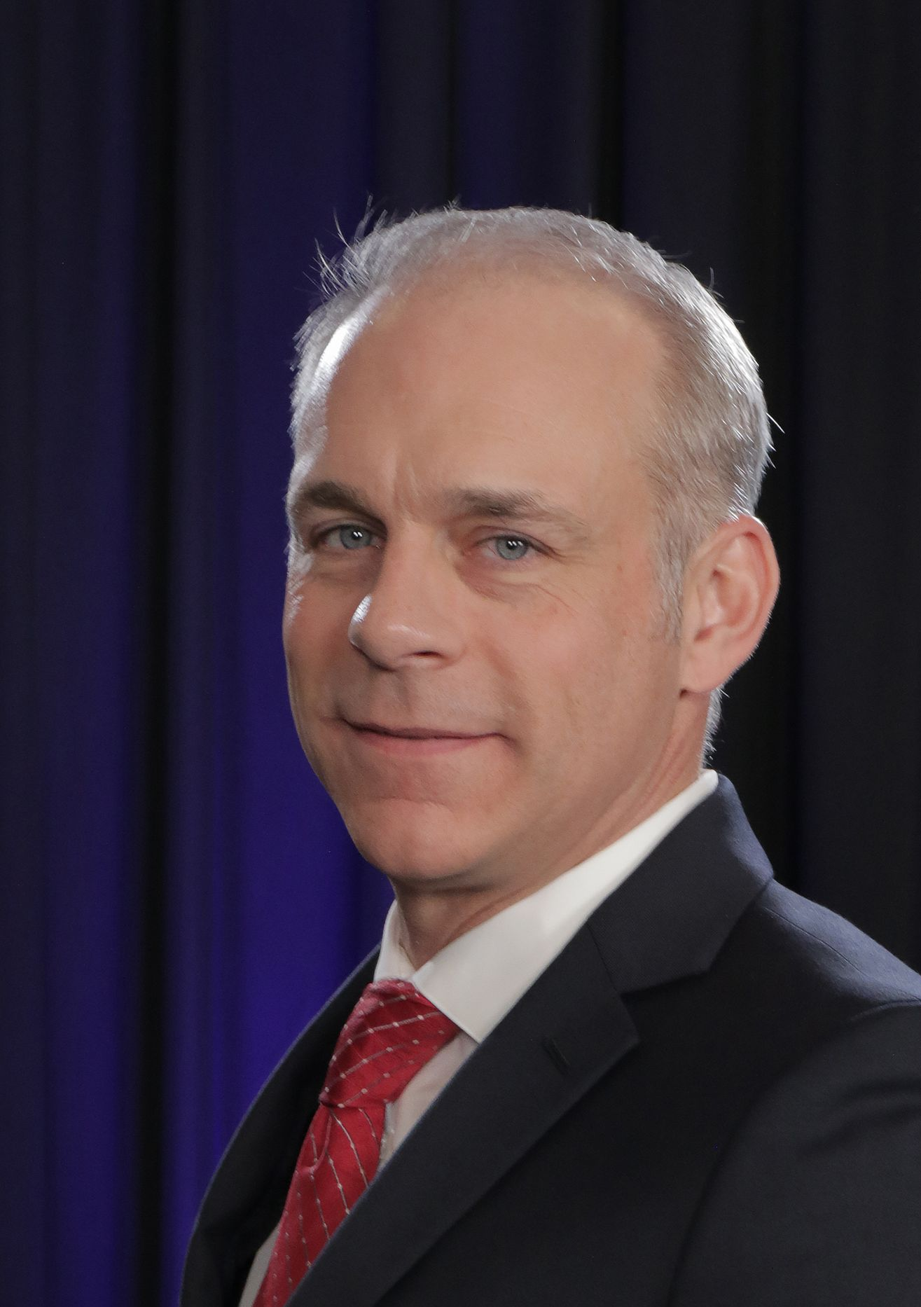 Smiling man in black coat, blue shirt, outdoors with mountains in background.