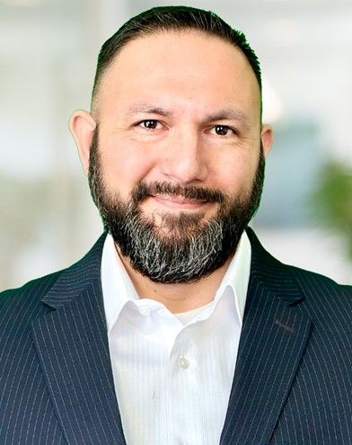 Man in suit, smiling, with beard and dark hair. Soft, neutral background.