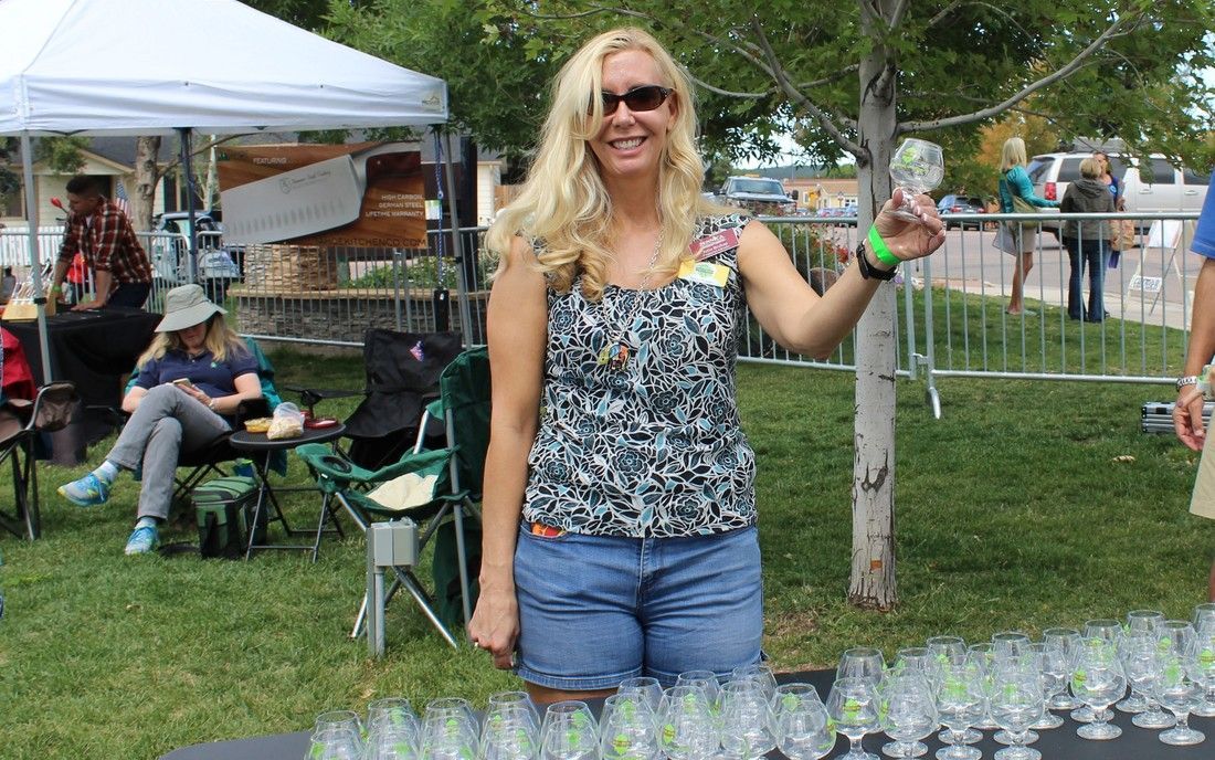 Woman holding glass, smiling at outdoor event. She wears shorts, tank top. Glasses line table, tents in background.
